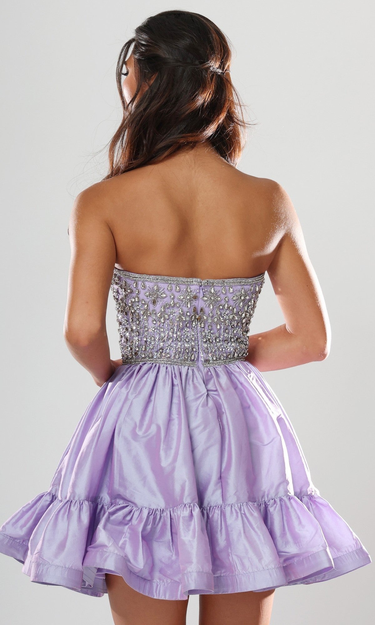 A woman stands with her back to the camera, modeling the Short Homecoming Dress: Jovani 52439, which features a strapless design with intricate beading on the bodice and a ruffled lilac skirt, set against a plain light background.